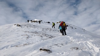 A group of people are hiking up a snowy mountain slope. They are dressed in winter gear, equipped with backpacks, and some are using trekking poles. The sky is overcast with clouds, and snow covers the ground, with patches of exposed rock.