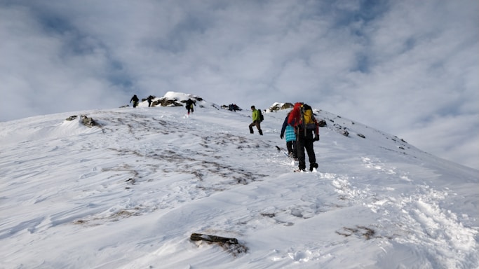 A group of people are hiking up a snowy mountain slope. They are dressed in winter gear, equipped with backpacks, and some are using trekking poles. The sky is overcast with clouds, and snow covers the ground, with patches of exposed rock.