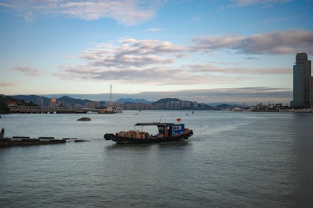 A small boat with various goods and a flag sails on a wide river or bay. The background features a cityscape with high-rise buildings and rolling hills under a partly cloudy sky. The water is calm and reflects the colors of the sky.