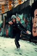 A person dressed in urban streetwear is striking a dynamic pose in a graffiti-covered alleyway. They are wearing a gray beanie, sunglasses, a dark jacket, and sneakers. The background features colorful graffiti art on a concrete wall and snow on the ground.
