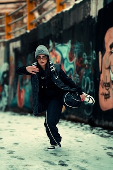 A person dressed in urban streetwear is striking a dynamic pose in a graffiti-covered alleyway. They are wearing a gray beanie, sunglasses, a dark jacket, and sneakers. The background features colorful graffiti art on a concrete wall and snow on the ground.