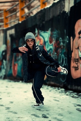 A person dressed in urban streetwear is striking a dynamic pose in a graffiti-covered alleyway. They are wearing a gray beanie, sunglasses, a dark jacket, and sneakers. The background features colorful graffiti art on a concrete wall and snow on the ground.