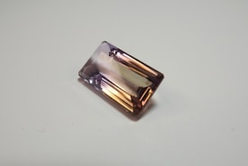 A rectangular, faceted gemstone with a transparent appearance and a gradient of colors ranging from light purple to golden brown. It is placed on a plain, light-colored surface.