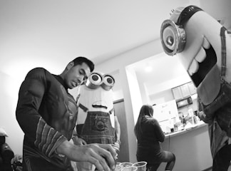 A person in a superhero costume is pouring drinks into cups, surrounded by two individuals dressed in large cartoon character costumes resembling Minions. In the background, a woman is seated at a kitchen counter in a casual setting.