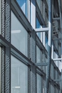 A modern architectural facade features large glass windows adorned with geometric patterns. Metal frames and beams create a grid-like structure, allowing ample light to penetrate. The design reflects urban sophistication and contemporary styling.