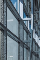 A modern architectural facade features large glass windows adorned with geometric patterns. Metal frames and beams create a grid-like structure, allowing ample light to penetrate. The design reflects urban sophistication and contemporary styling.