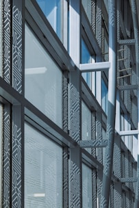 A modern architectural facade features large glass windows adorned with geometric patterns. Metal frames and beams create a grid-like structure, allowing ample light to penetrate. The design reflects urban sophistication and contemporary styling.