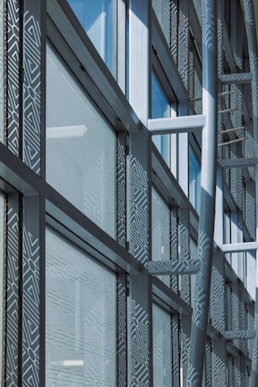A modern architectural facade features large glass windows adorned with geometric patterns. Metal frames and beams create a grid-like structure, allowing ample light to penetrate. The design reflects urban sophistication and contemporary styling.