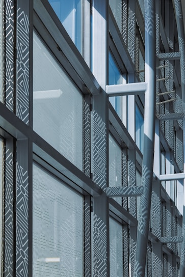 A modern architectural facade features large glass windows adorned with geometric patterns. Metal frames and beams create a grid-like structure, allowing ample light to penetrate. The design reflects urban sophistication and contemporary styling.