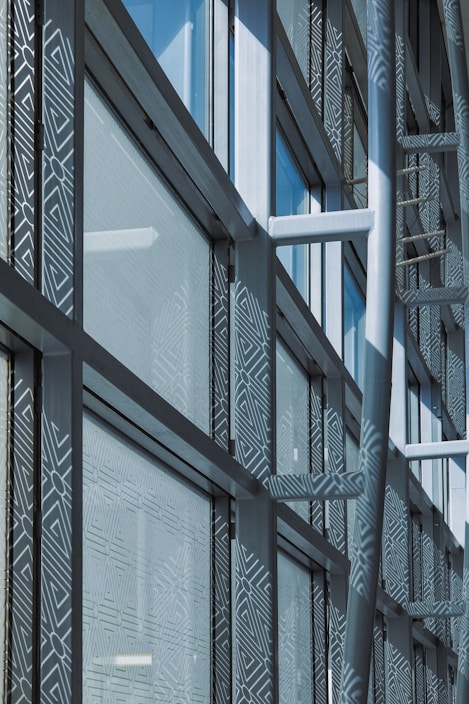 A modern architectural facade features large glass windows adorned with geometric patterns. Metal frames and beams create a grid-like structure, allowing ample light to penetrate. The design reflects urban sophistication and contemporary styling.