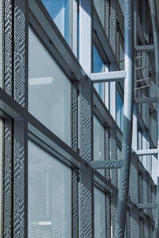 A modern architectural facade features large glass windows adorned with geometric patterns. Metal frames and beams create a grid-like structure, allowing ample light to penetrate. The design reflects urban sophistication and contemporary styling.