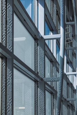A modern architectural facade features large glass windows adorned with geometric patterns. Metal frames and beams create a grid-like structure, allowing ample light to penetrate. The design reflects urban sophistication and contemporary styling.