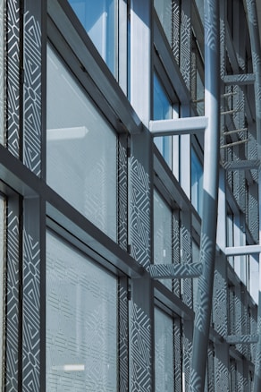 A modern architectural facade features large glass windows adorned with geometric patterns. Metal frames and beams create a grid-like structure, allowing ample light to penetrate. The design reflects urban sophistication and contemporary styling.