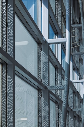 A modern architectural facade features large glass windows adorned with geometric patterns. Metal frames and beams create a grid-like structure, allowing ample light to penetrate. The design reflects urban sophistication and contemporary styling.