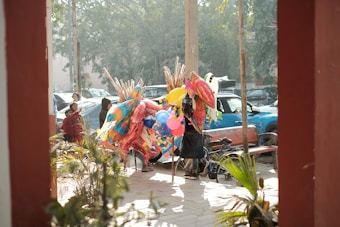 A street vendor is surrounded by colorful inflatable toys and balloons, with several people nearby. The scene is set in an outdoor market or street setting with parked cars and trees in the background. Sunlight filters through the trees, casting shadows on the ground and creating a lively atmosphere.