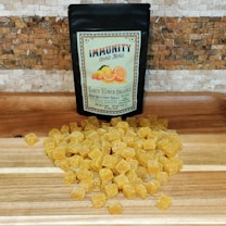 A black package labeled 'Immunity Orange-Mango' from Earth Flower Organics is placed on a wooden surface. In front of the package, numerous small yellow cubes with a sugary coating are spread across the surface. The backdrop consists of a textured, mosaic-style wall.