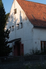 A house with white textured walls and a steeply pitched roof covered in reddish-brown shingles. The house has small windows and a door with a wooden awning. There are trees and bushes surrounding the structure, and a tall wooden fence in the foreground. The light creates shadows of the nearby trees on the house's facade.