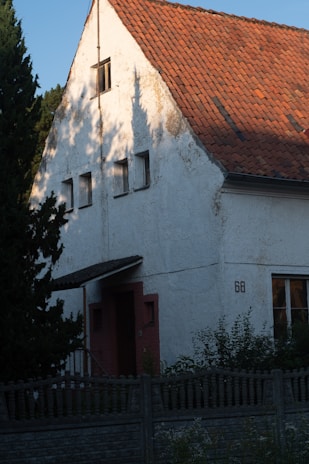 A house with white textured walls and a steeply pitched roof covered in reddish-brown shingles. The house has small windows and a door with a wooden awning. There are trees and bushes surrounding the structure, and a tall wooden fence in the foreground. The light creates shadows of the nearby trees on the house's facade.