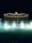 A grand hotel building illuminated at night with arches and a central dome, accompanied by a large fountain displaying choreographed water jets and mist in front.