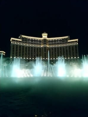 A grand hotel building illuminated at night with arches and a central dome, accompanied by a large fountain displaying choreographed water jets and mist in front.