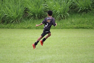 A child wearing a black soccer uniform and red-orange cleats, running on a lush green field. Tall grass surrounds the area, indicating an outdoor setting, possibly a park or sports field.
