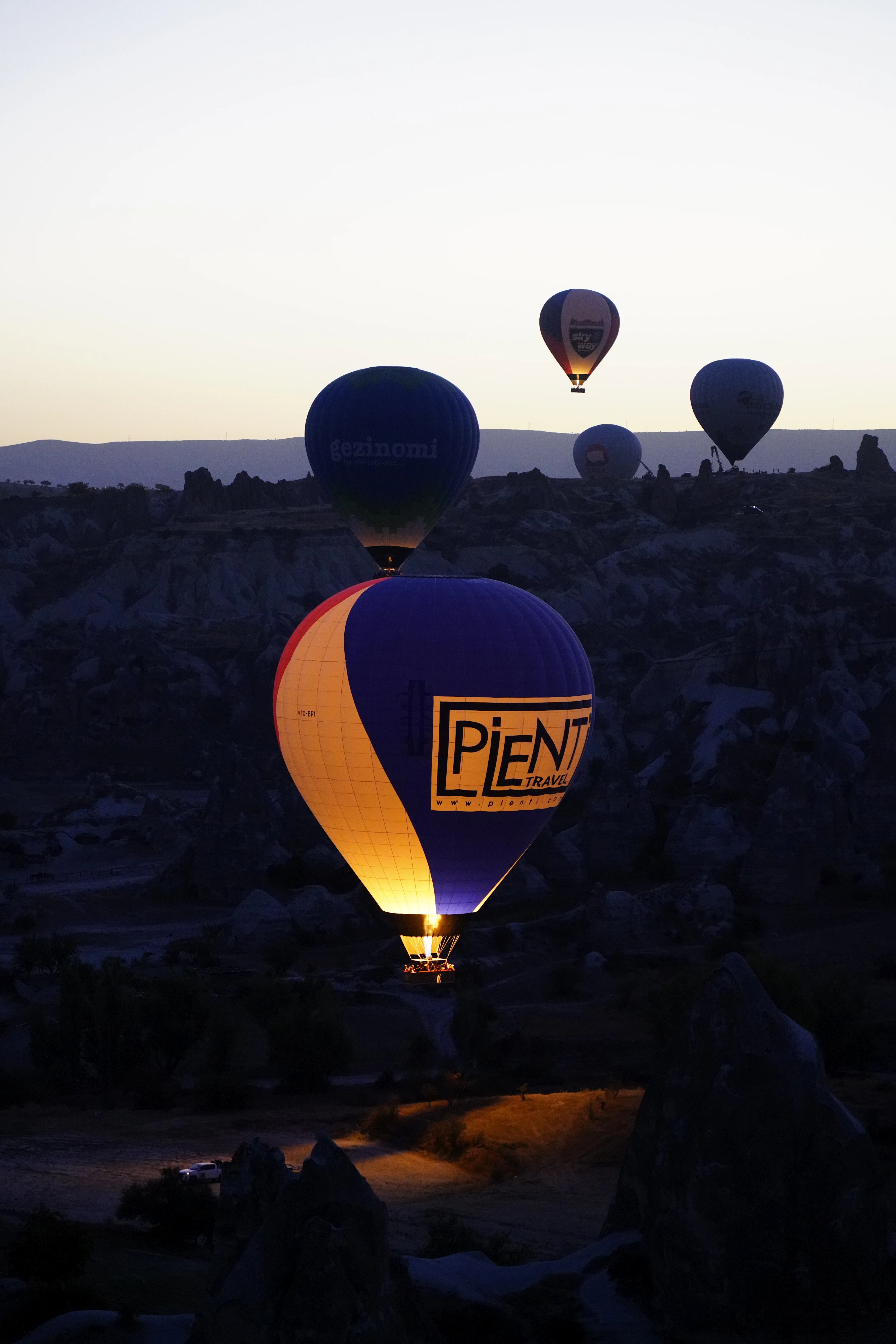 A hot air balloon glowing from its burner flame against the dark valley at dawn, more balloons rising behind it.