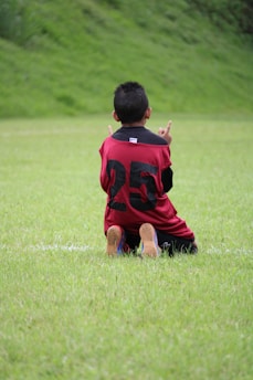 A child is kneeling on a grassy field, wearing a red and black sports jersey with the number 25 on the back. The child has short, dark hair and is raising both hands, with fingers pointing upward. The background features lush green grass and a slightly blurred green landscape, suggesting a natural setting like a park or sports field.