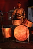 A metallic sculpture depicting a drummer with a drum set stands illuminated in warm light. The sculpture features exaggerated facial features and limbs, with drumsticks in hand. The background reveals a dimly lit building with arched windows.