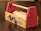 A small wooden toy toolbox with bright red sides and a wooden dowel handle. The box displays a ruler along the top edge and has a blue and a yellow knob on the front panel. The words 'TOOL BOX' are printed in blue on the side.