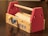 A small wooden toy toolbox with bright red sides and a wooden dowel handle. The box displays a ruler along the top edge and has a blue and a yellow knob on the front panel. The words 'TOOL BOX' are printed in blue on the side.