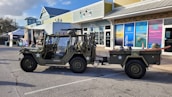 A military-style off-road vehicle is parked in a commercial area, towing a small trailer equipped with a surfboard. The area features storefronts with colorful advertisements and a variety of outdoor displays. The vehicle is painted in an olive green typical of military vehicles and adorned with white stars and identification numbers. Surrounding the scene are a few small businesses with modern architecture.
