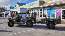 A military-style off-road vehicle is parked in a commercial area, towing a small trailer equipped with a surfboard. The area features storefronts with colorful advertisements and a variety of outdoor displays. The vehicle is painted in an olive green typical of military vehicles and adorned with white stars and identification numbers. Surrounding the scene are a few small businesses with modern architecture.