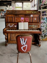 A wooden upright piano is covered with colorful graffiti and stickers, positioned in an abandoned or unfinished room with concrete floors and graffiti-covered walls. An open sheet of music sits on the piano stand, while a single chair with a red cushion and white graffiti is placed in front of it. The room is filled with natural light streaming through large windows in the background.