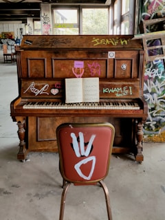 A wooden upright piano is covered with colorful graffiti and stickers, positioned in an abandoned or unfinished room with concrete floors and graffiti-covered walls. An open sheet of music sits on the piano stand, while a single chair with a red cushion and white graffiti is placed in front of it. The room is filled with natural light streaming through large windows in the background.