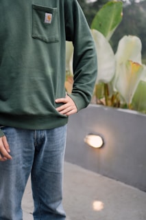 A person is dressed in a green sweatshirt with a logo on the pocket and a pair of blue jeans. The person is standing with one hand on their hip. The background includes large green leaves and a smooth concrete surface illuminated by a light fixture.
