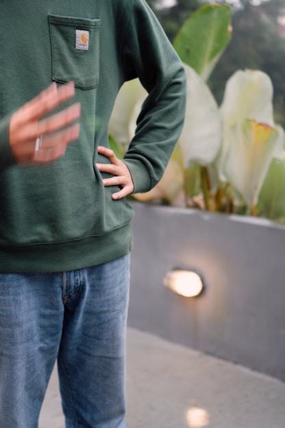 A person is wearing a dark green sweatshirt with a chest pocket, paired with light blue jeans. The background features large green leaves and a gray wall with a small light fixture.