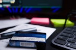 A close-up of a desk containing a tobacco product box with a health warning label, placed on top of printed documents. Nearby, there are neon-colored sticky notes, a pen, and part of a keyboard visible. The background includes a blurred computer screen.