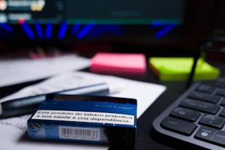 A close-up of a desk containing a tobacco product box with a health warning label, placed on top of printed documents. Nearby, there are neon-colored sticky notes, a pen, and part of a keyboard visible. The background includes a blurred computer screen.