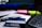 A close-up of a desk containing a tobacco product box with a health warning label, placed on top of printed documents. Nearby, there are neon-colored sticky notes, a pen, and part of a keyboard visible. The background includes a blurred computer screen.