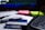 A close-up of a desk containing a tobacco product box with a health warning label, placed on top of printed documents. Nearby, there are neon-colored sticky notes, a pen, and part of a keyboard visible. The background includes a blurred computer screen.