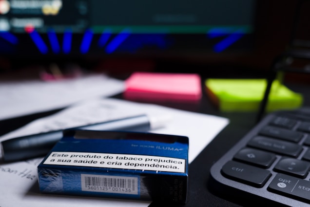 A close-up of a desk containing a tobacco product box with a health warning label, placed on top of printed documents. Nearby, there are neon-colored sticky notes, a pen, and part of a keyboard visible. The background includes a blurred computer screen.
