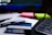 A close-up of a desk containing a tobacco product box with a health warning label, placed on top of printed documents. Nearby, there are neon-colored sticky notes, a pen, and part of a keyboard visible. The background includes a blurred computer screen.