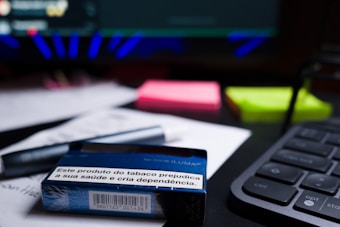 A close-up of a desk containing a tobacco product box with a health warning label, placed on top of printed documents. Nearby, there are neon-colored sticky notes, a pen, and part of a keyboard visible. The background includes a blurred computer screen.