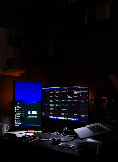 A tech workspace with two computer monitors displaying a variety of applications. The left monitor shows a communication platform, while the right one is filled with a multi-column interface for managing tasks or projects. A keyboard, mouse, headphones, and a laptop are on the desk, along with a smartphone and various stationery items. Dim ambient lighting creates a focused and somewhat secluded atmosphere.