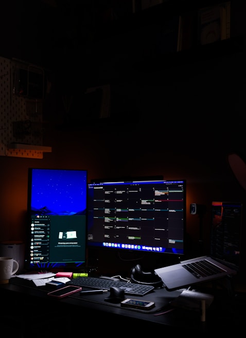 A tech workspace with two computer monitors displaying a variety of applications. The left monitor shows a communication platform, while the right one is filled with a multi-column interface for managing tasks or projects. A keyboard, mouse, headphones, and a laptop are on the desk, along with a smartphone and various stationery items. Dim ambient lighting creates a focused and somewhat secluded atmosphere.