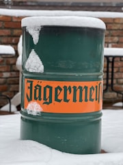 A green metal barrel with an orange label displaying bold, black text is partially covered in snow. The background consists of a brick wall and patio furniture, indicating an outdoor setting.