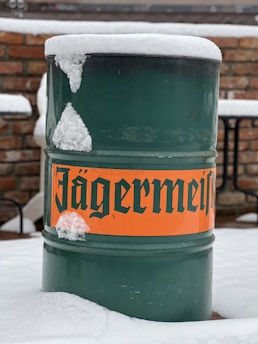 A green metal barrel with an orange label displaying bold, black text is partially covered in snow. The background consists of a brick wall and patio furniture, indicating an outdoor setting.