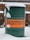 A green metal barrel with an orange label displaying bold, black text is partially covered in snow. The background consists of a brick wall and patio furniture, indicating an outdoor setting.