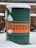 A green metal barrel with an orange label displaying bold, black text is partially covered in snow. The background consists of a brick wall and patio furniture, indicating an outdoor setting.