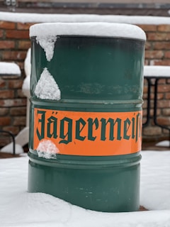 A green metal barrel with an orange label displaying bold, black text is partially covered in snow. The background consists of a brick wall and patio furniture, indicating an outdoor setting.